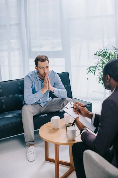 Young man sitting on couch and talking to african american counselor — Stock Photo