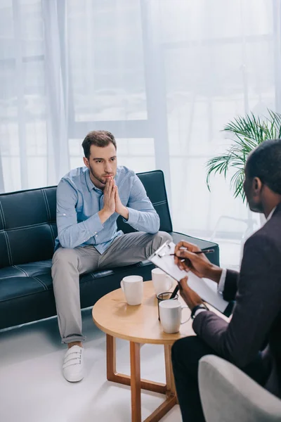 Joven sentado en el sofá y hablando con el consejero afroamericano - foto de stock