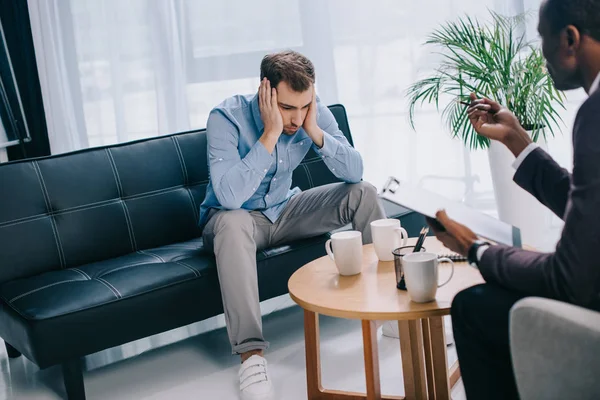 Upset young man sitting on couch and psychiatrist with clipboard — Stock Photo