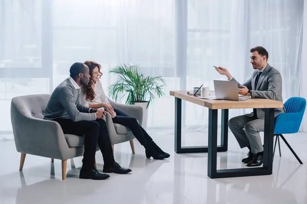 Couple Interracial assis dans des fauteuils et agent immobilier à table avec ordinateur portable — Photo de stock