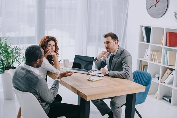 Agente inmobiliario sentado en la mesa con el ordenador portátil y hablando con los clientes - foto de stock