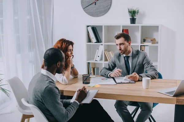 Agente inmobiliario con pareja interracial sentada en la mesa en la oficina - foto de stock