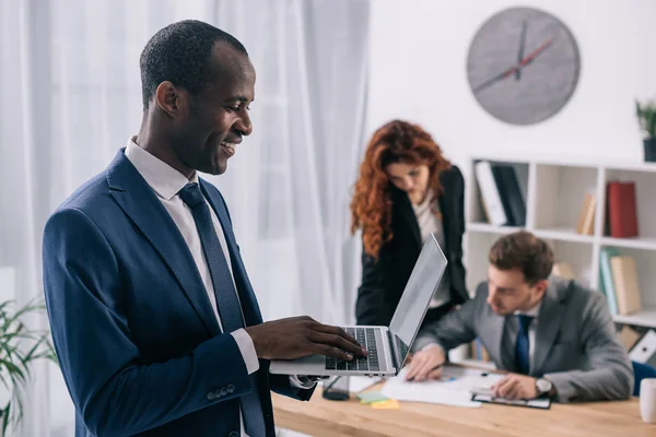 Hombre de negocios africano sonriente con portátil en las manos y dos colegas de negocios trabajando en la mesa - foto de stock