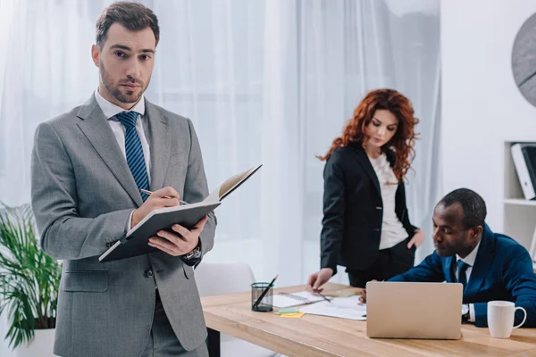 Joven hombre de negocios con colegas haciendo papeleo en la mesa con el portátil - foto de stock