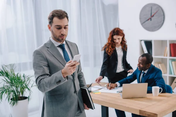 Young stylish businessman with smartphone and two business partners doing paperwork at table — Stock Photo