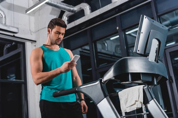 Deportista escuchando música con auriculares y smartphone mientras entrena en la cinta de correr en el gimnasio — Stock Photo