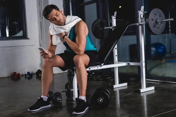 Deportista sudoroso con toalla de descanso y el uso de teléfono inteligente en el gimnasio - foto de stock