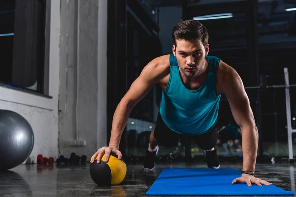 Muscular sportsman doing push ups with medicine ball on yoga mat in sports center — Stock Photo