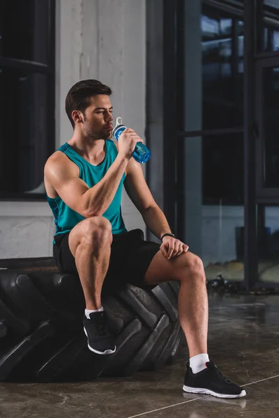 Sportsman sitting on tire and drinking water after cross training in gym — Stock Photo