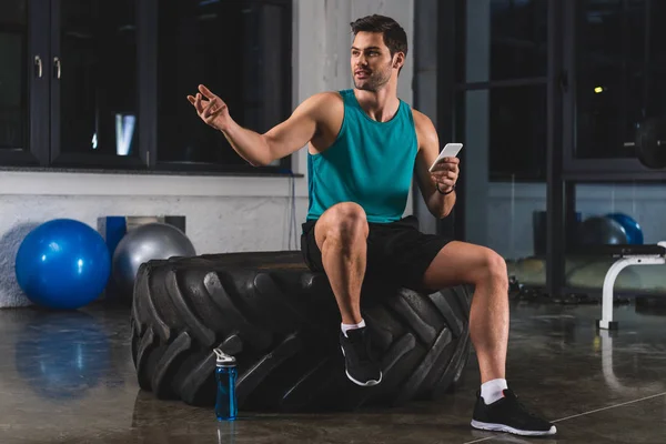 Sportsman sitting on tire and using smartphone in gym — Stock Photo