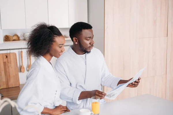 Africain américain lecture de journal et petite amie souriante dans la cuisine — Photo de stock