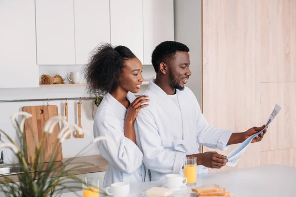 Улыбающийся африканский американец читает газету, а его девушка обнимает его на кухне. — стоковое фото