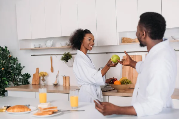 Smiling woman giving apple to boyfriend in kitchen — Stock Photo