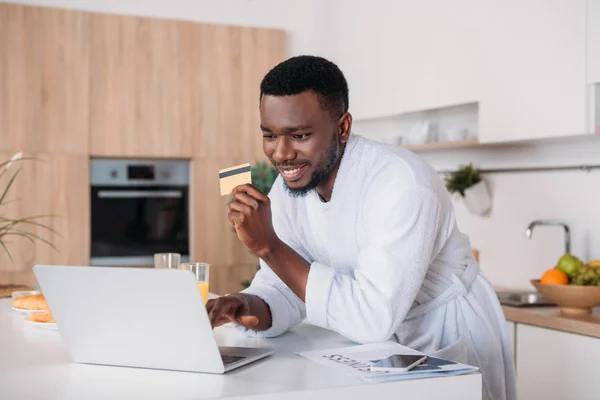 Lächelnder Mann beim Online-Shopping und steht mit Kreditkarte in der Küche — Stockfoto