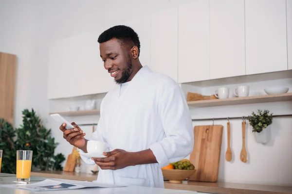 Africano americano com copo de café digitando no smartphone na cozinha — Fotografia de Stock