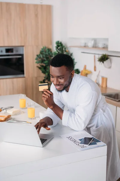 Smiling man doing online shopping and standing with credit card in kitchen — Stock Photo