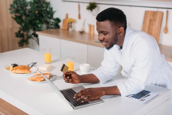 African american man doing online shopping and standing with credit card in kitchen — Stock Photo