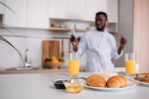 Vista de perto de croissants no prato, suco de laranja, compotas e manteiga com o jovem em fones de ouvido no fundo embaçado — Fotografia de Stock