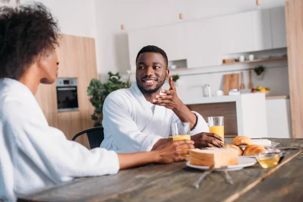 Sonriente hombre afroamericano hablando con su novia mientras está sentado en la mesa con el desayuno - foto de stock