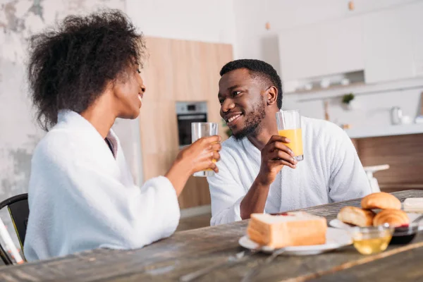 Jovem casal segurando óculos com suco e sentado à mesa com torradas e croissants — Fotografia de Stock