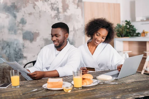 Hombre afroamericano desayunando con su novia haciendo compras en línea en la cocina - foto de stock