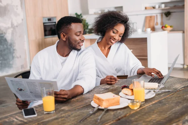 Young man with newspaper and smiling girlfriend with credit card doing online shopping — Stock Photo