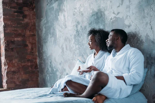 Vista lateral de una pareja sonriente sentada en la cama con tazas de café - foto de stock