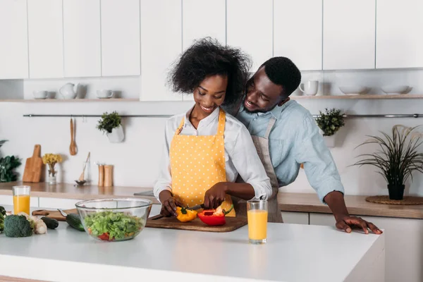 Smiling man hugging girlfriend while she cutting pepper on board in kitchen — Stock Photo