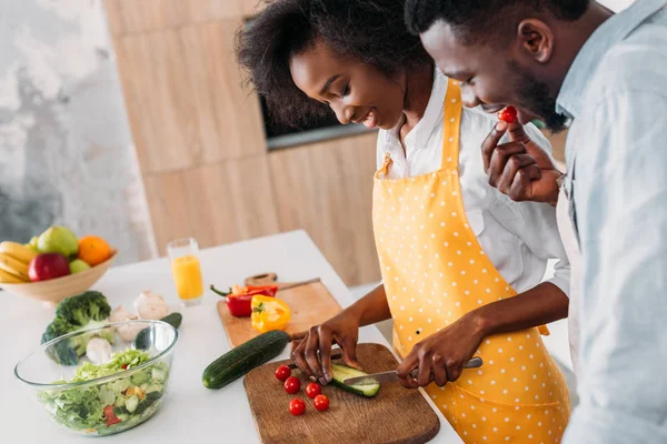 Young woman cutting cucumber on board and boyfriend eating cherry tomato — Stock Photo