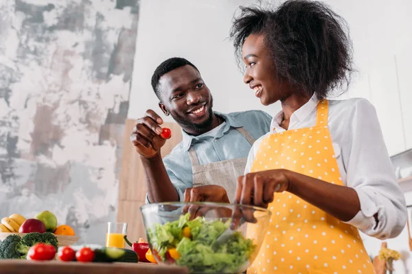 Низький кут зору молодої жінки, змішуючи салат в мисці і чоловік тримає вишневий помідор в руці — стокове фото