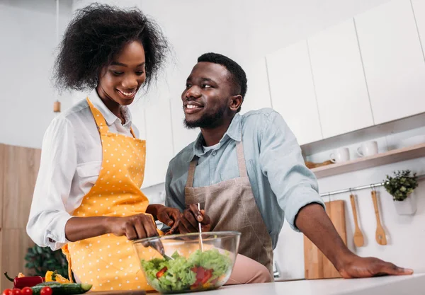 Low angle view of smiling woman mixing salad in bowl and sitting on table with boyfriend standing near — Stock Photo