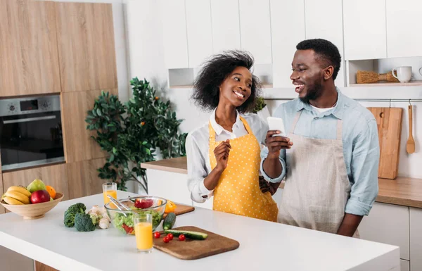 Afrikanisch-amerikanisches Paar in Schürzen steht mit Essen am Tisch und benutzt Smartphone in der Küche — Stockfoto