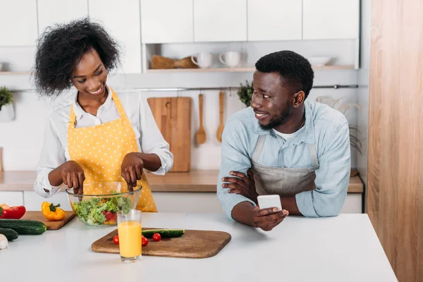 Femme mélangeant salade dans un bol à table et petit ami debout avec smartphone à la main — Photo de stock