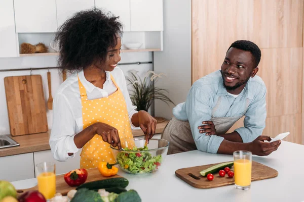 Femme souriante mélangeant salade dans un bol à table et petit ami debout avec smartphone à la main — Photo de stock