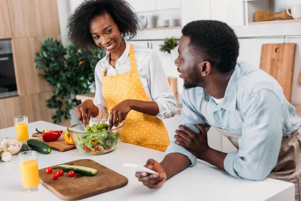 Young woman mixing salad in bowl at table and boyfriend standing with smartphone in hand — Stock Photo