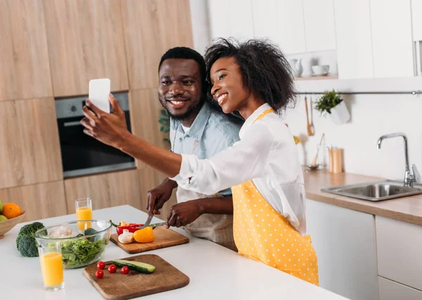 Sorrindo jovem casal tomando selfie enquanto cortando legumes a bordo à mesa — Fotografia de Stock