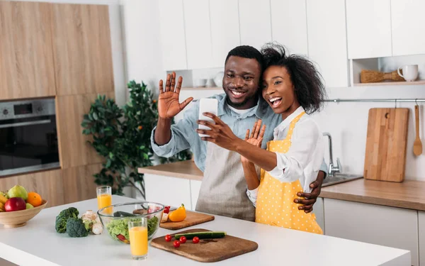 Улыбающаяся африканская американская пара в фартуках делает селфи на кухне — стоковое фото