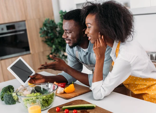 Junges Paar schaut sich Rezept auf digitalem Tablet an, während es mit Gemüse am Tisch steht — Stockfoto