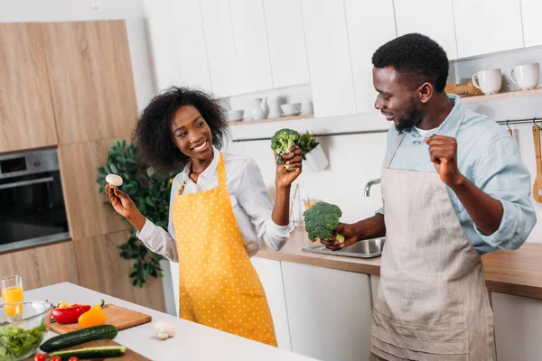 Couple souriant dans des tabliers tenant champignons et brocoli — Photo de stock