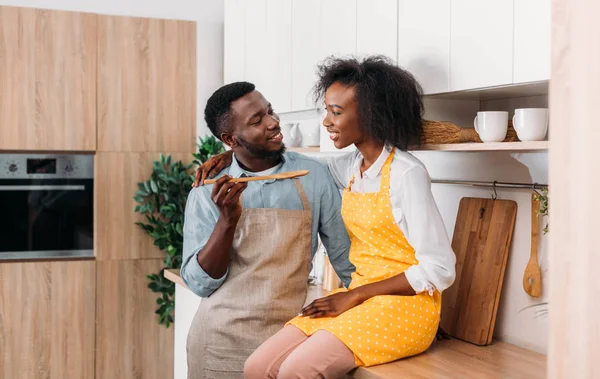 Jeune couple souriant dans des tabliers dans la cuisine avec une cuillère en bois — Photo de stock