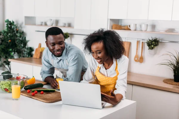 Sonriente pareja joven usando portátil en la mesa con verduras en la cocina - foto de stock
