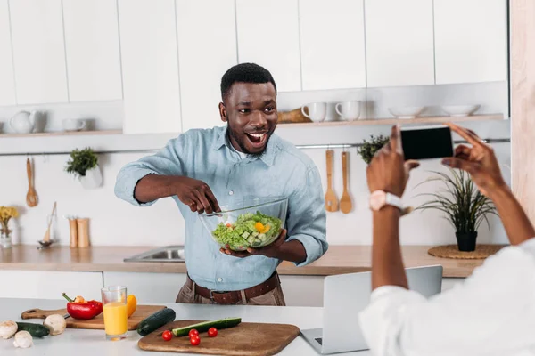 Cropped image of woman taking photo of boyfriend holding bowl with salad — Stock Photo