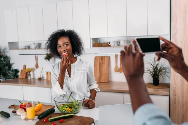 Vista ritagliata dell'uomo che scatta foto di una giovane donna afroamericana che mangia pomodoro ciliegia — Foto stock