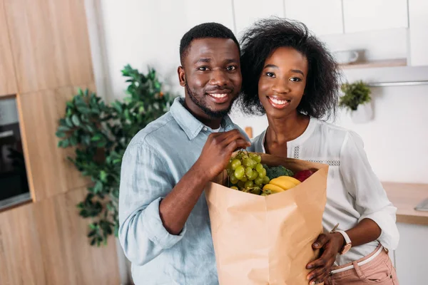 Sorrindo casal afro-americano segurando saco de papel com frutas na cozinha — Fotografia de Stock