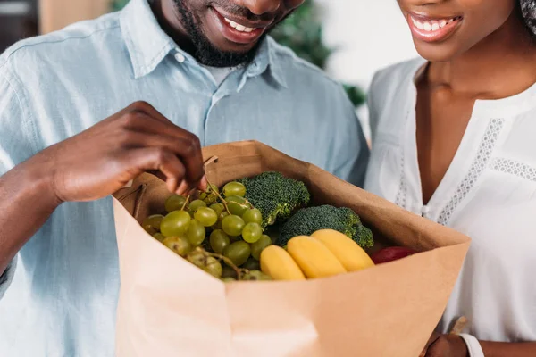 Cropped view of young couple holding paper bag with grapes, bananas and broccoli — Stock Photo