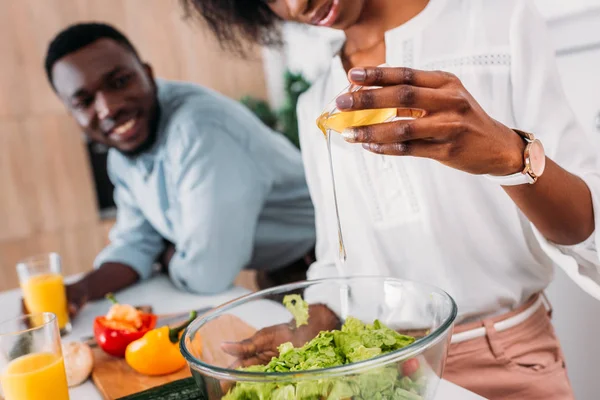 Vista ritagliata della donna afro-americana che aggiunge olio in insalata mentre sorride fidanzato in piedi accanto — Foto stock