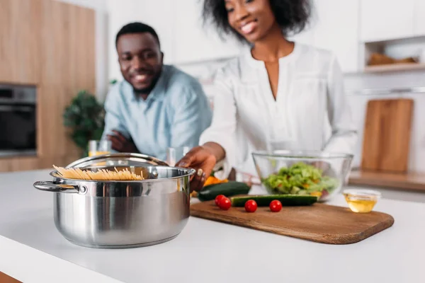 Vista de cerca de la cacerola con pasta y verduras en la mesa con una pareja sonriente detrás - foto de stock