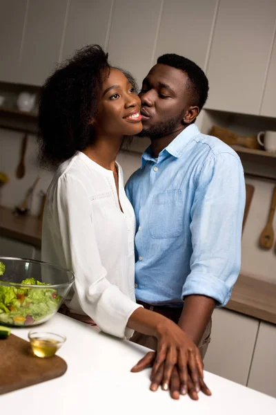 Мужчина целует молодую улыбающуюся женщину на кухне — стоковое фото