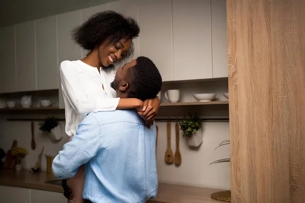 Joven afroamericano hombre levantando sonriente novia en cocina - foto de stock