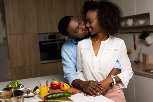 Smiling african american couple hugging near table with vegetables and saucepan — Stock Photo